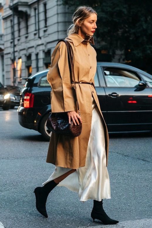 Stylish woman in trench coat walking on city street, carrying a brown handbag, urban fashion.
