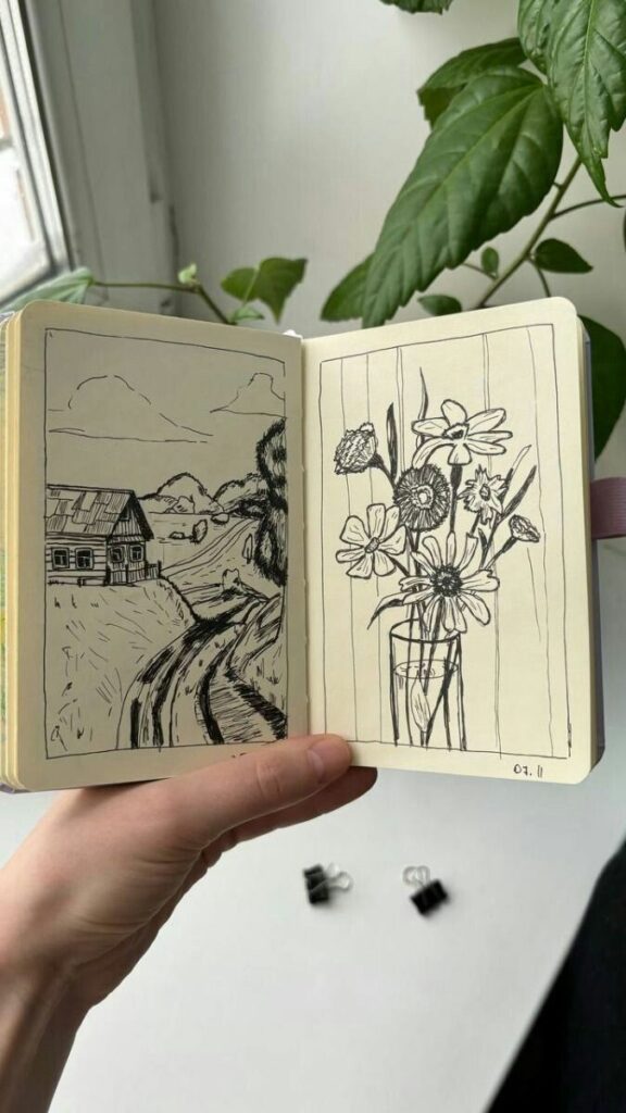 Open sketchbook with hand-drawn landscape and flower illustrations held near a window with green plants.