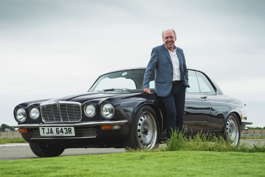Man in a blue blazer poses with a classic black vintage car on a cloudy day, showcasing timeless style.