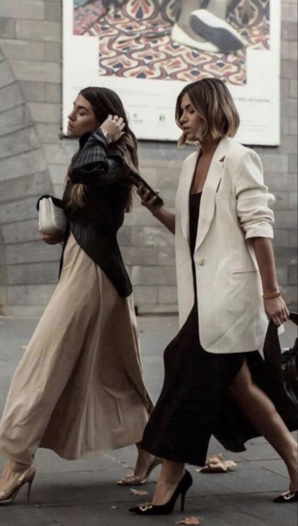 Two women walking stylishly in urban setting, wearing chic long skirts and elegant blazers.