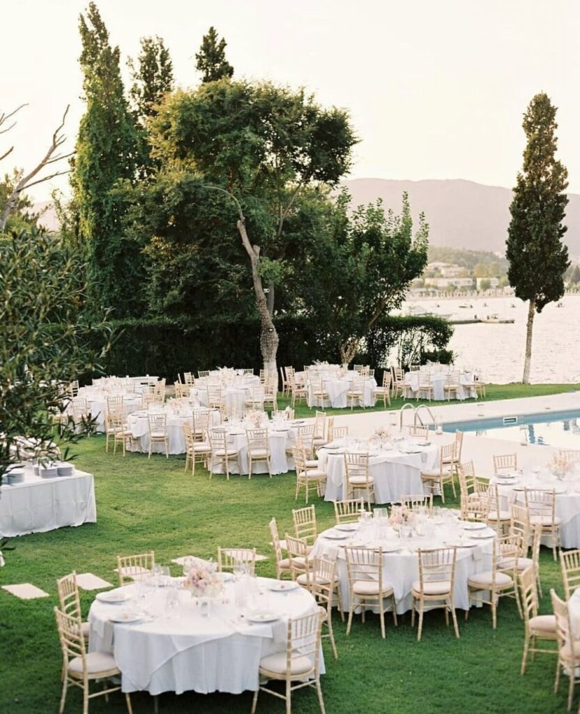 Elegant outdoor wedding reception with round tables, white linens, and wooden chairs beside a pool and scenic view.