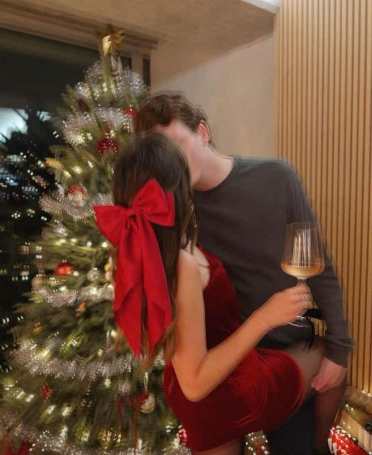 Couple sharing a holiday kiss by a decorated Christmas tree with festive lights, holding a glass of white wine.