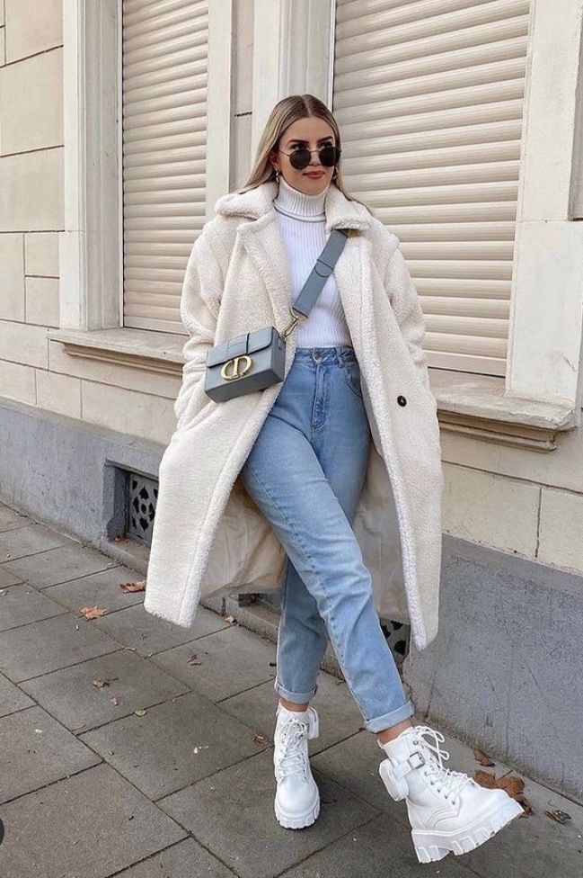 Woman in stylish winter outfit: white coat, jeans, boots, sunglasses, and shoulder bag, posing outdoors. Fashionable street style.
