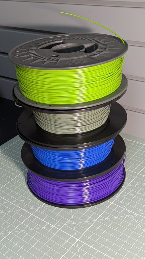 2e3b3459ae7c98e37ba13d0ca5d82602 Stack of colorful 3D printer filament spools: green, gray, blue, and purple, on a grid mat. | Sky Rye Design Stack of colorful 3D printer filament spools: green, gray, blue, and purple, on a grid mat.