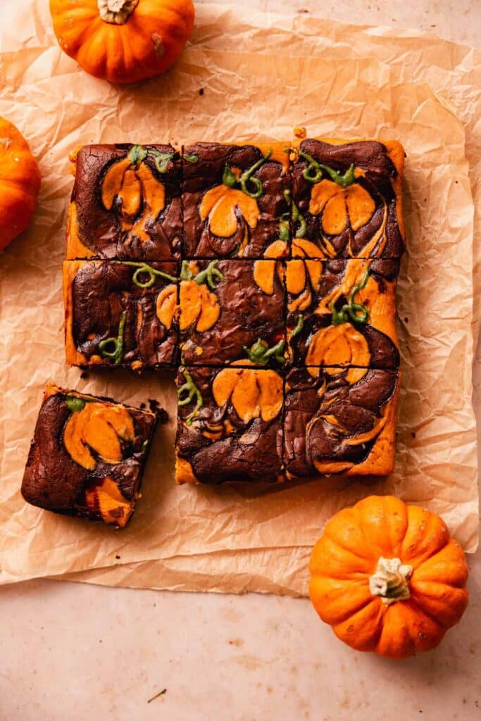 Pumpkin brownies with chocolate swirl, cut into squares, surrounded by small pumpkins on parchment paper.