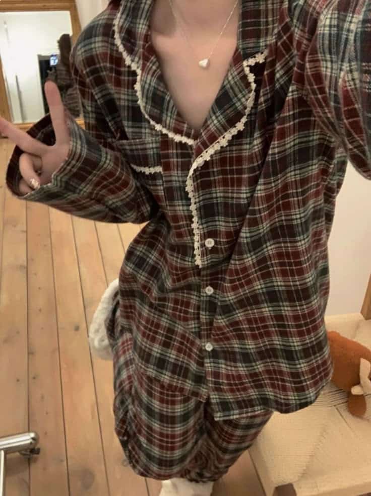 Person in cozy plaid pajamas making a peace sign, standing on wooden floor near a chair and teddy bear.