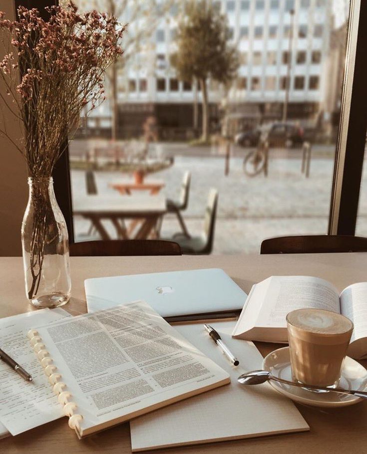 Cozy cafe workspace with laptop, open books, latte, and flowers by a window, perfect for study or remote work.