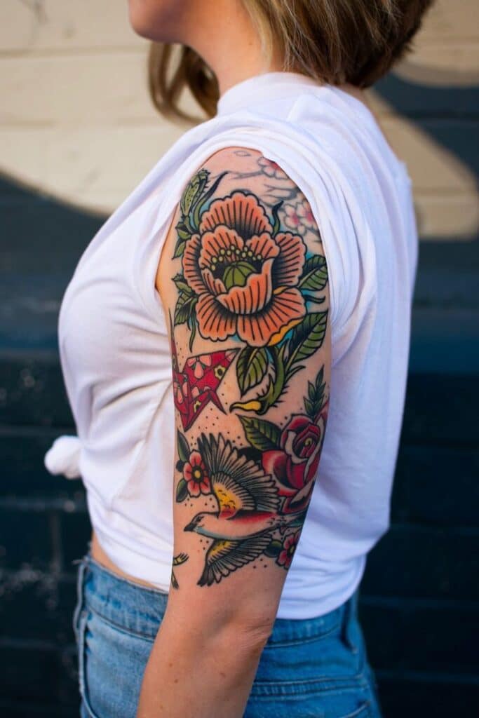 Colorful floral tattoo sleeve with birds and roses on a woman's arm, vibrant art in a casual outfit.