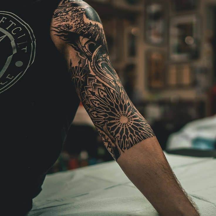 Intricate black sleeve tattoo featuring mandala and geometric patterns on an arm in a tattoo studio setting.