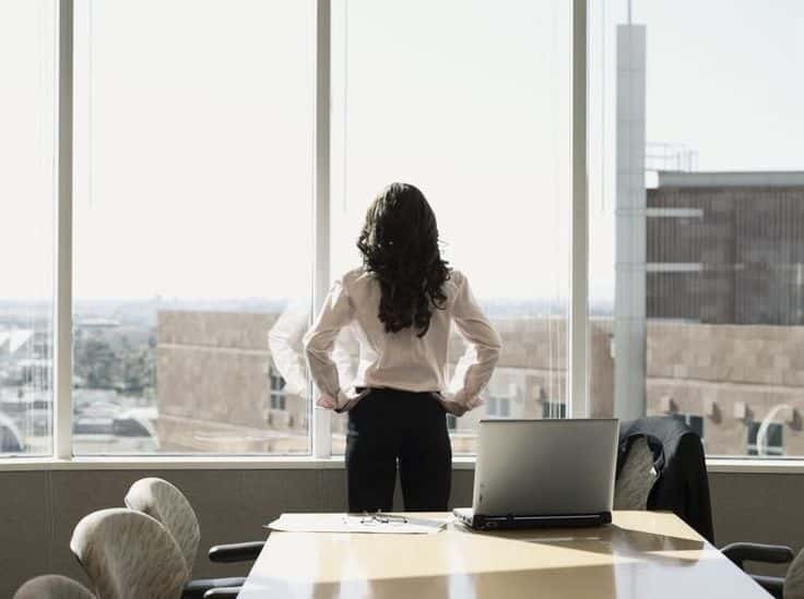Businesswoman looking out office window, standing with hands on hips beside a conference table and laptop.