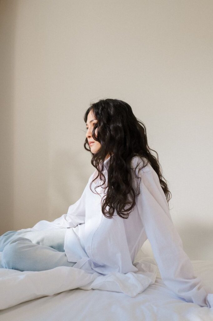 Woman with long curly hair in white shirt sitting on bed in a serene, minimalist room.