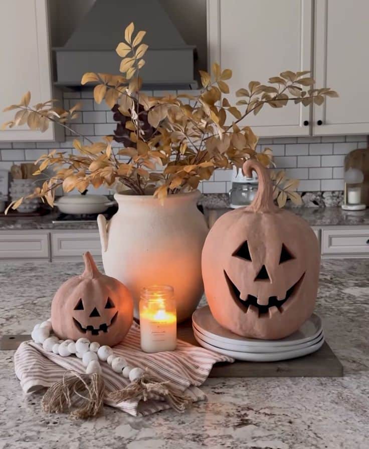 Halloween decor with two jack-o'-lanterns, candle, and autumn leaves on a kitchen counter. Cozy fall ambiance.