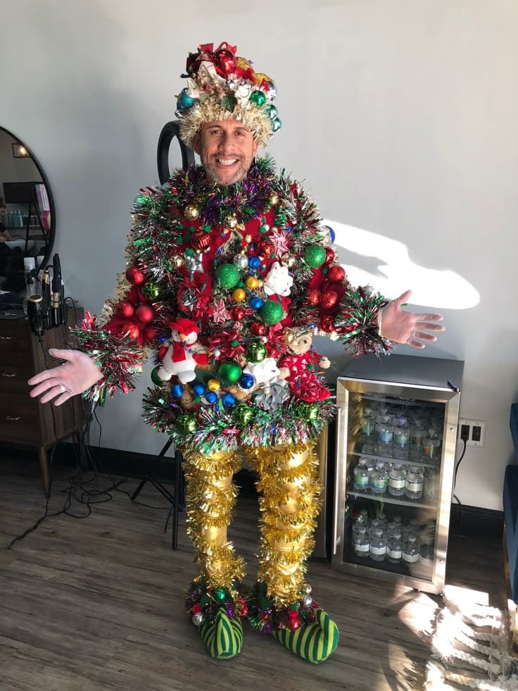 Man dressed as festive Christmas tree with ornaments and tinsel, standing indoors smiling with arms open.