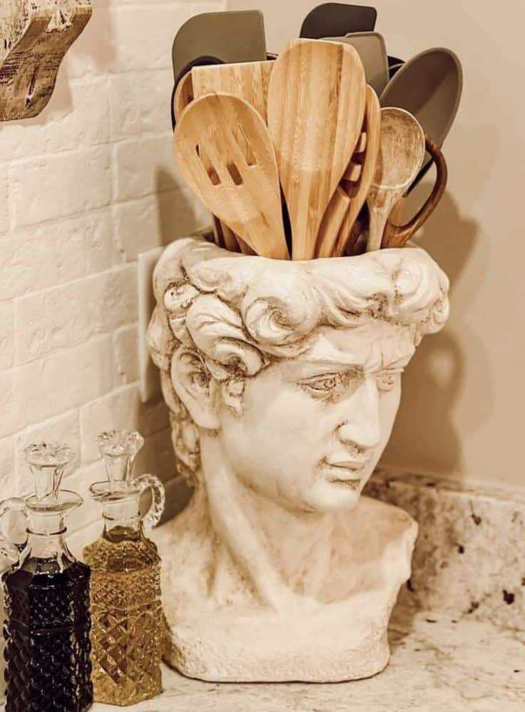 Classical statue head utensil holder with wooden kitchen tools on a marble countertop.