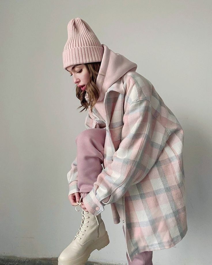 Woman in plaid coat, pink hoodie, and beanie lacing boots. Stylish winter fashion outfit.