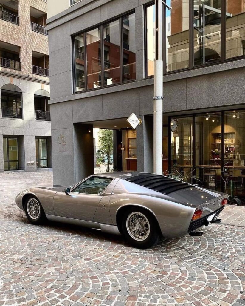 Classic silver sports car parked on cobblestone street outside modern building.
