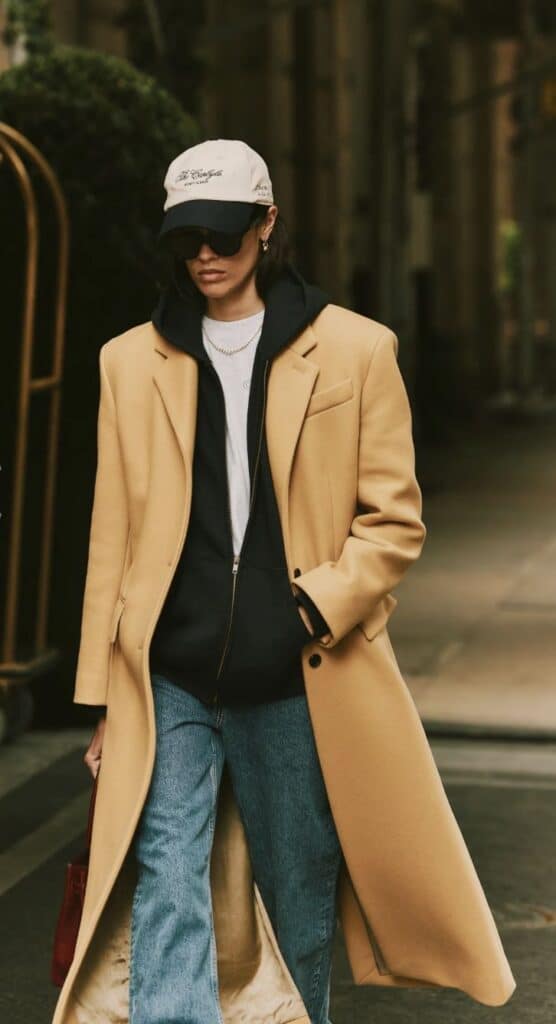Stylish person in camel coat, black hoodie, and jeans, wearing a cap and sunglasses, walking confidently in an urban setting.