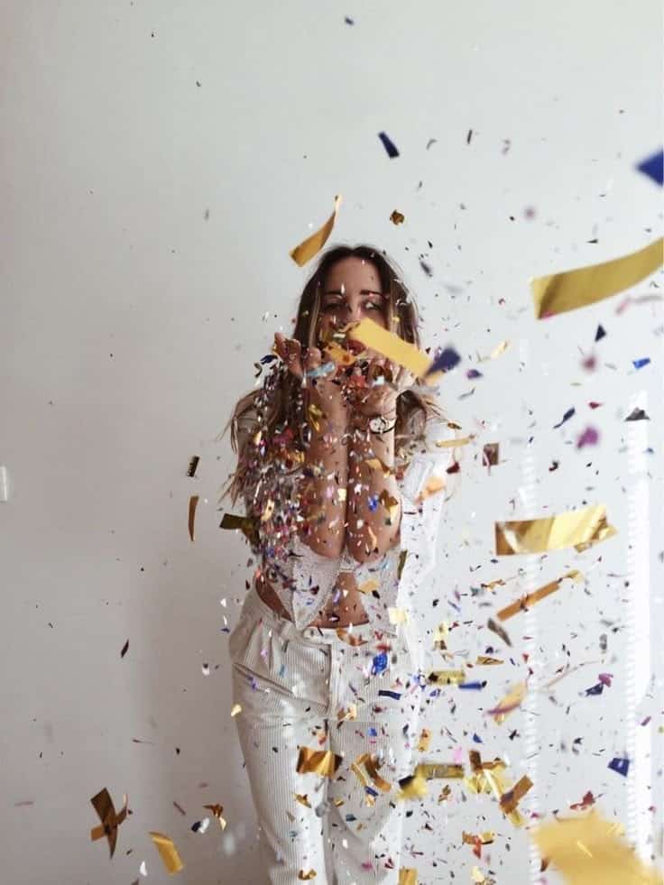 Person celebrating with vibrant confetti flying in the air, wearing white pants and a light top, against a plain background.