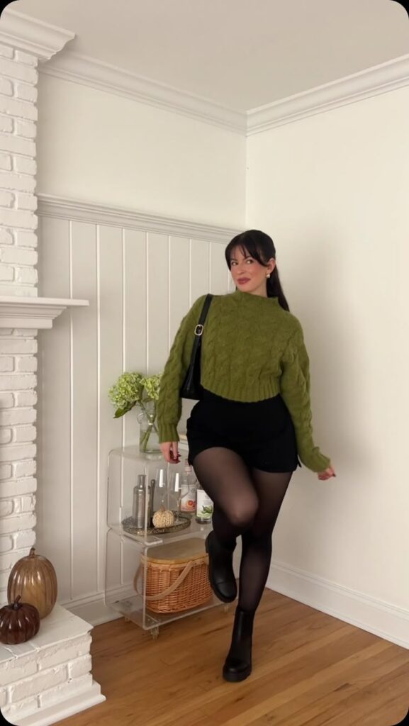Woman in a green sweater and black shorts posing indoors with decor and plants. Stylish fall fashion.