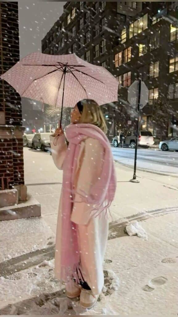 Person with pink umbrella and scarf in snowy cityscape at night.