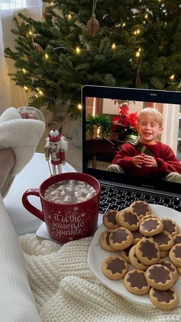 Cozy Christmas scene with hot chocolate, cookies, and a laptop playing a holiday movie by a lit tree.