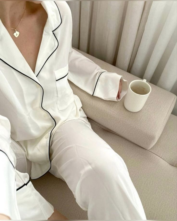Person in white pajamas with black piping holding a mug, relaxing on a beige couch near curtains. Cozy morning vibe.