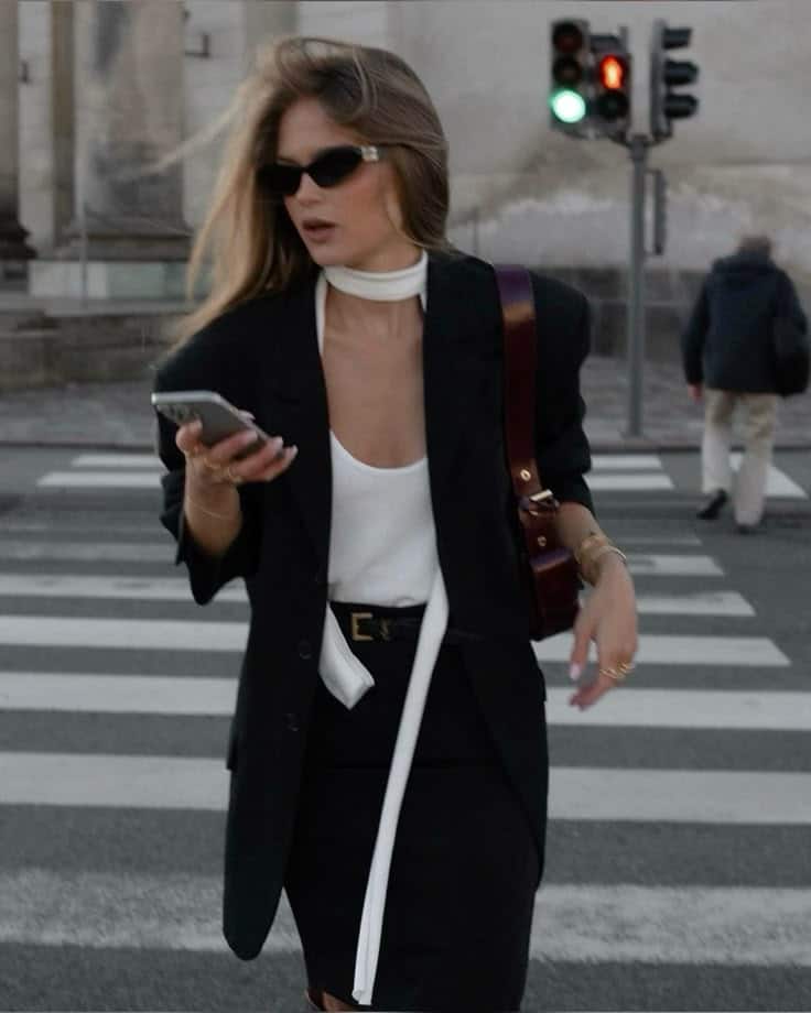 Fashionable woman in sunglasses crossing the street while checking her phone, chic urban style with black blazer.