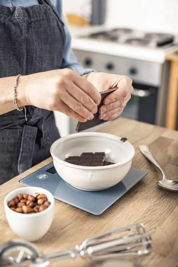 Person breaking chocolate into a bowl for melting, with hazelnuts and whisk nearby on a wooden kitchen counter.