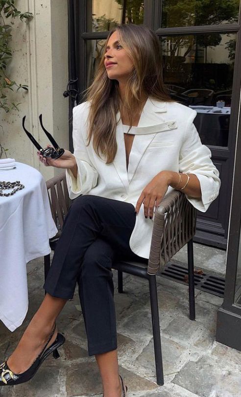 Woman in stylish white blazer and black pants sitting at an outdoor café, holding sunglasses.