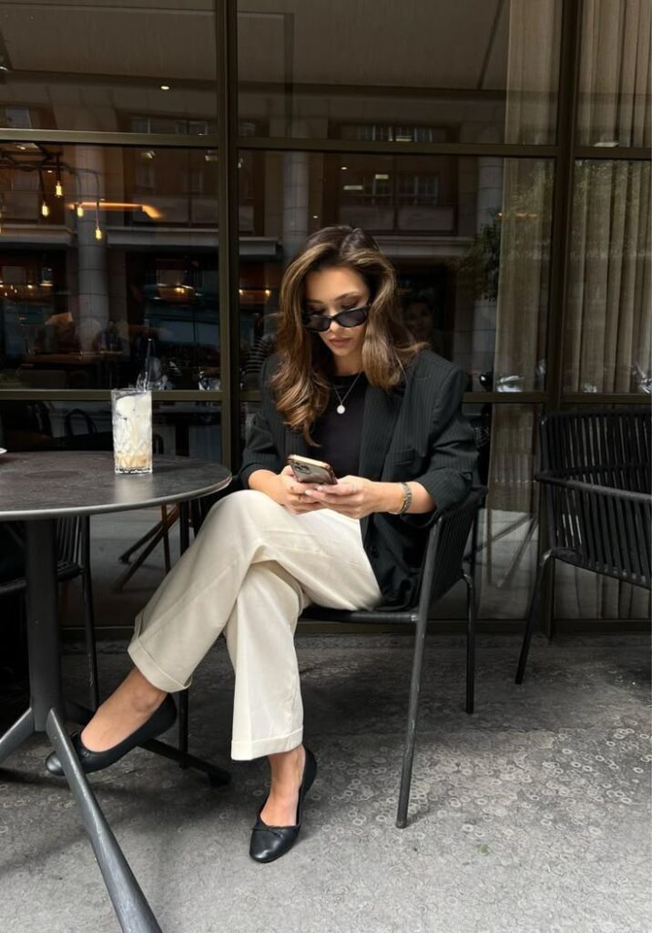 Woman in sunglasses seated at outdoor cafe, checking phone with drink on table. Urban setting, casual style.