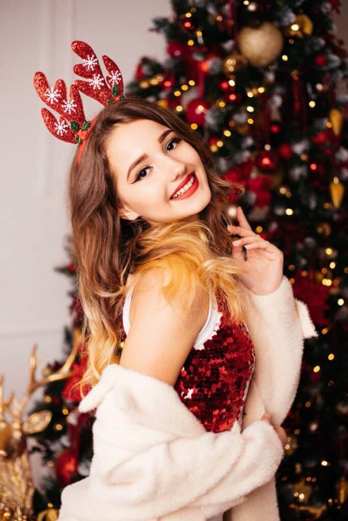 Smiling woman in festive antlers in front of a Christmas tree with red decorations.