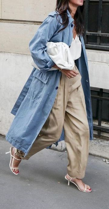Woman in stylish blue trench coat and beige pants holding a white clutch, walking confidently in high-heeled sandals.