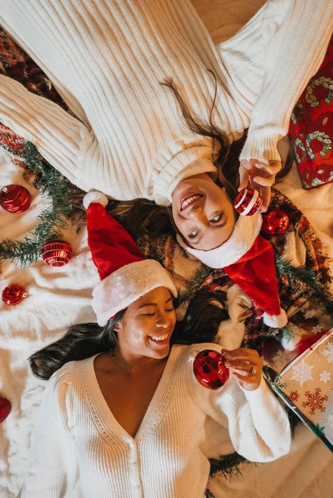 Two women wearing Santa hats and white sweaters lie on a bed with Christmas ornaments, smiling and festive. Christmas party