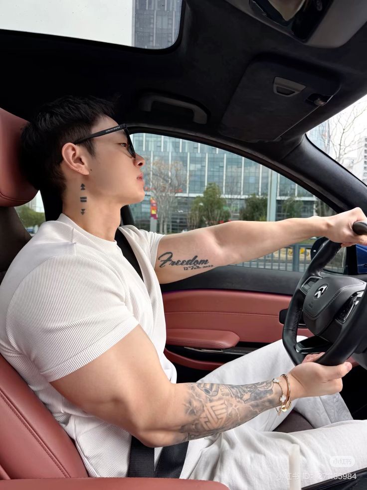 Man with tattoos driving a car, wearing a white shirt and sunglasses, focused on the road.