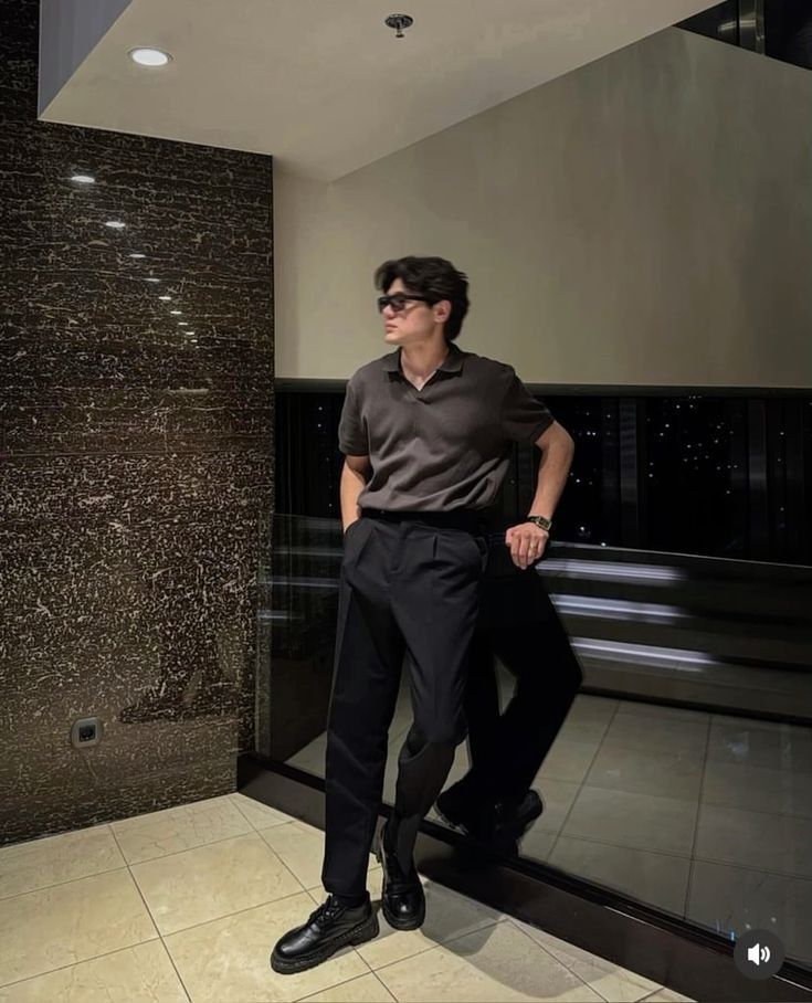 Stylish person in sunglasses and dark outfit, leaning against a wall in an elegant interior setting.