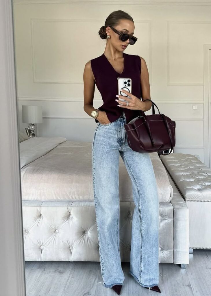 Woman in stylish outfit with sunglasses, high-waisted jeans, and burgundy top, standing in chic bedroom with phone and bag.