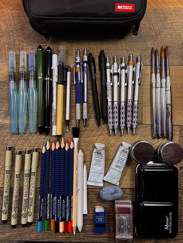 Art supplies neatly organized: pencils, pens, brushes, watercolors, and erasers on a wooden table with a black case.