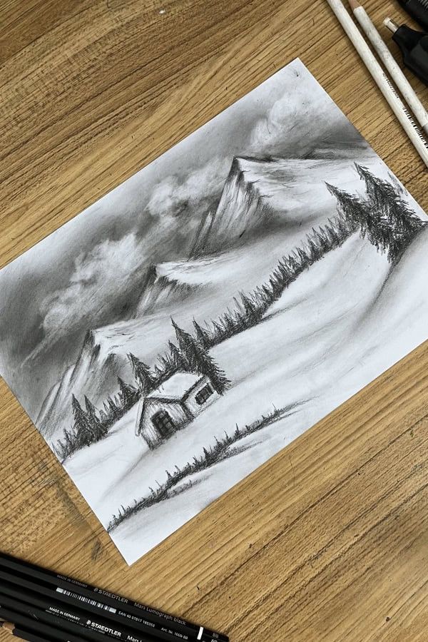 Charcoal sketch of a snowy mountain landscape with a cabin and pine trees on a wooden desk.