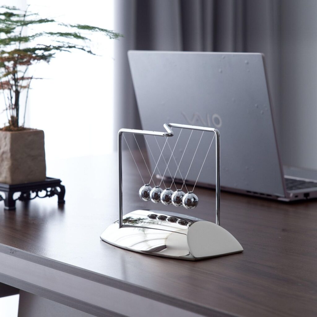 3f01320bfdb053b8f5276731b4e8915d Newton's cradle on a desk with laptop and plant, symbolizing balance and motion in a modern office setting. | Sky Rye Design Newton's cradle on a desk with laptop and plant, symbolizing balance and motion in a modern office setting.
