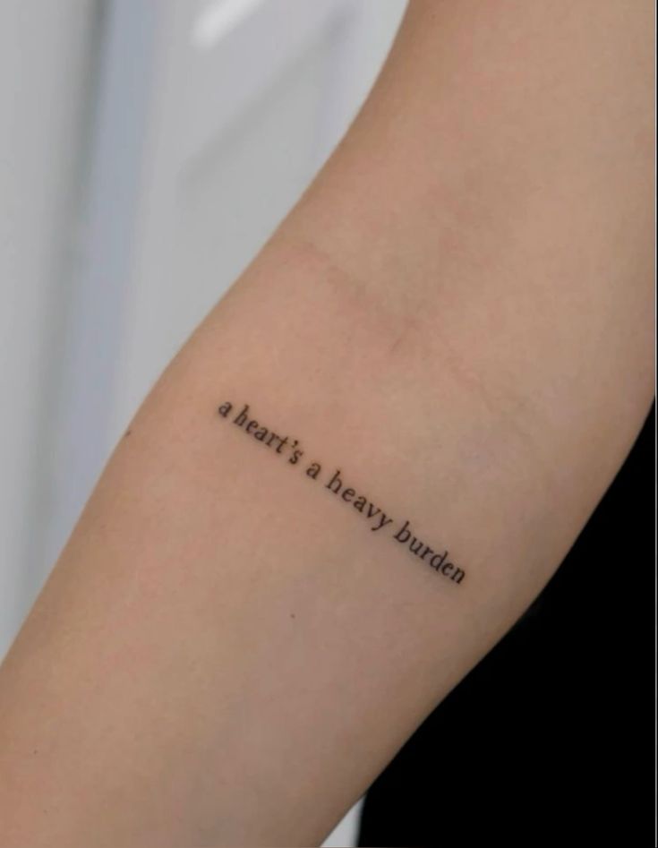 Minimalist arm tattoo reads a heart's a heavy burden in black ink.