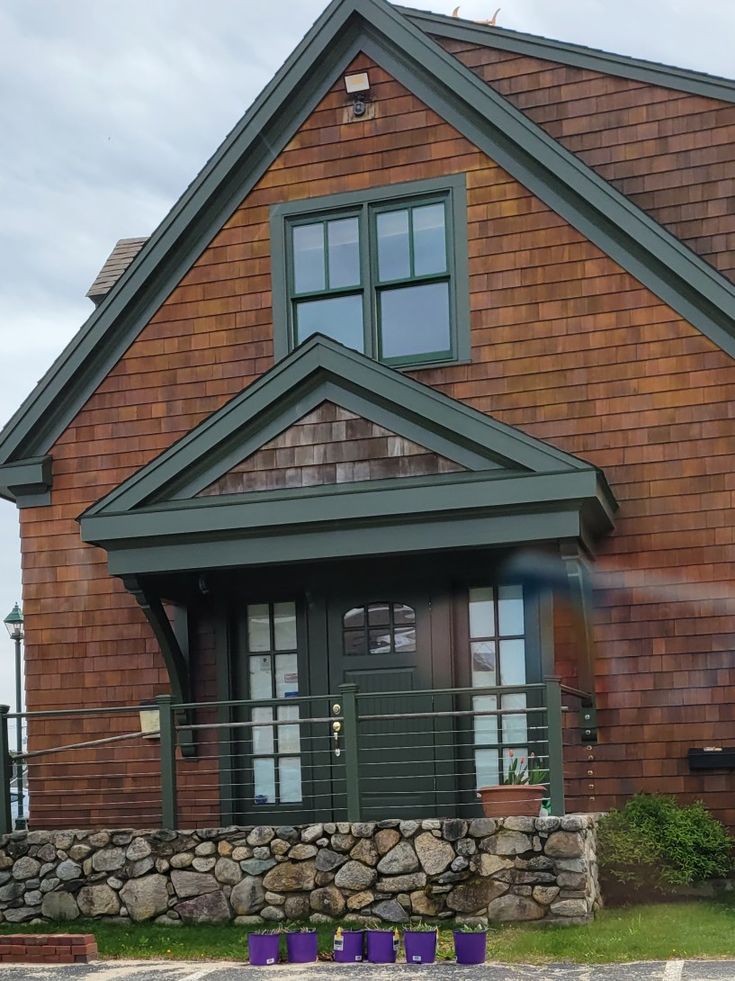 Cottage-style house with wooden shingles, stone foundation, and potted plants in purple containers by the entrance.