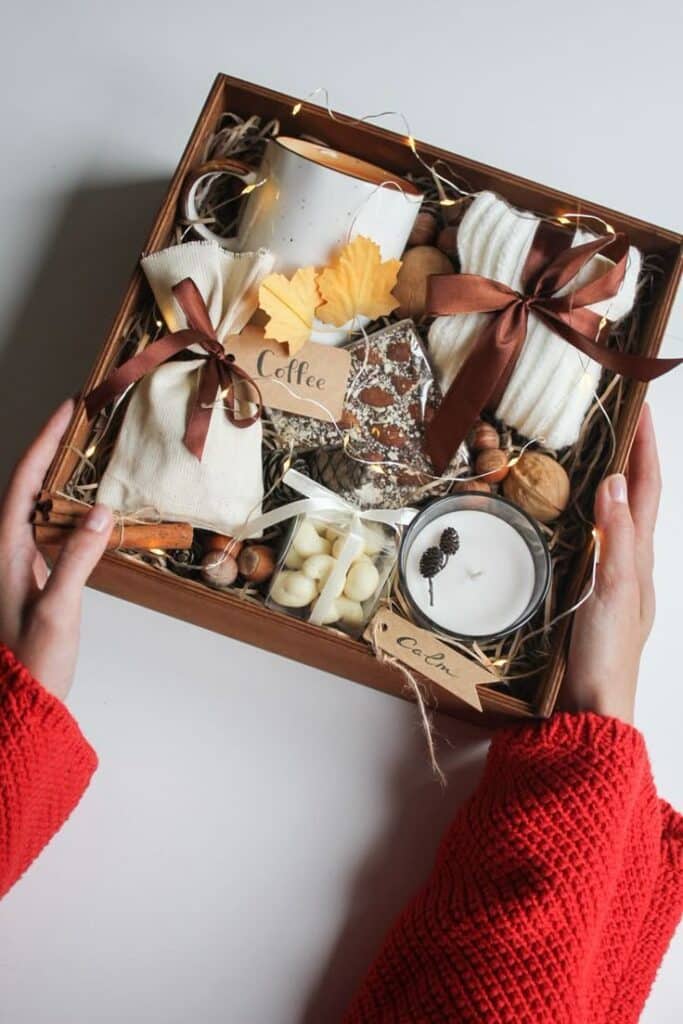 _ 4 Gift box with coffee, snacks, candle, and autumn decor, held in hands wearing a red sweater. Cozy and warm presentation. | Sky Rye Design Gift box with coffee, snacks, candle, and autumn decor, held in hands wearing a red sweater. Cozy and warm presentation.