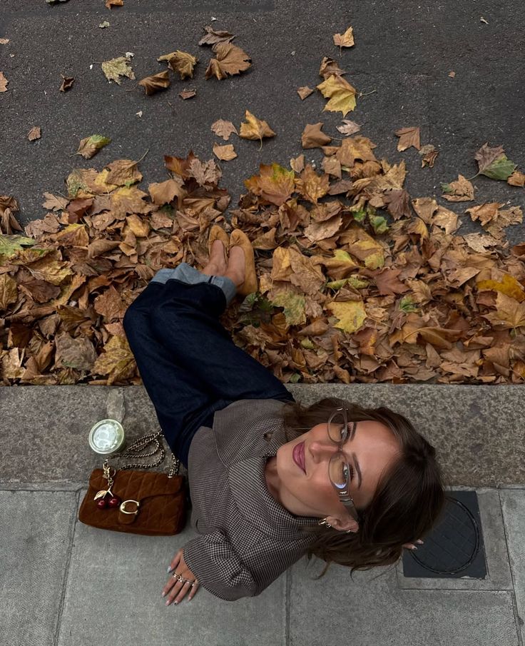 Woman sitting on a curb with autumn leaves, wearing glasses and holding a drink beside a brown handbag.