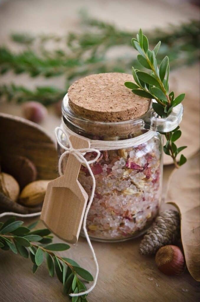 Glass jar of floral bath salts with wooden scoop, decorated with greenery, on a rustic table.