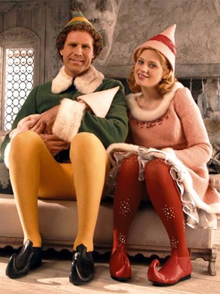 Two people in festive elf costumes sitting side by side on a bench, smiling in a cozy, holiday-themed setting.