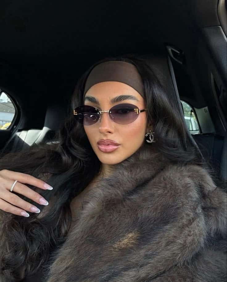 Woman in sunglasses with long hair and fur coat sitting in a car, posing with elegance and confidence.
