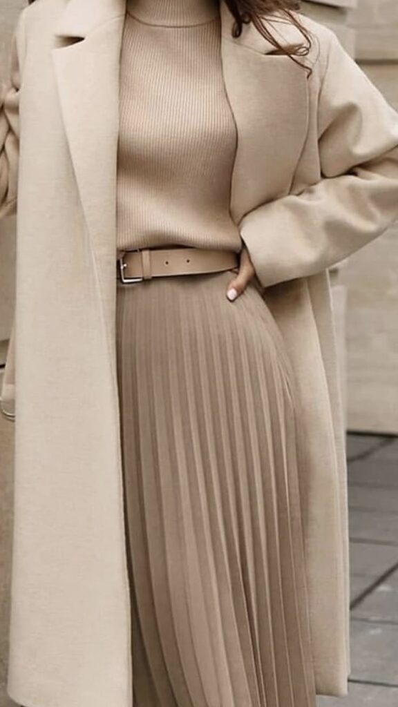 Woman in stylish beige outfit with long coat, ribbed sweater, pleated skirt, and matching belt, showcasing elegant fashion.