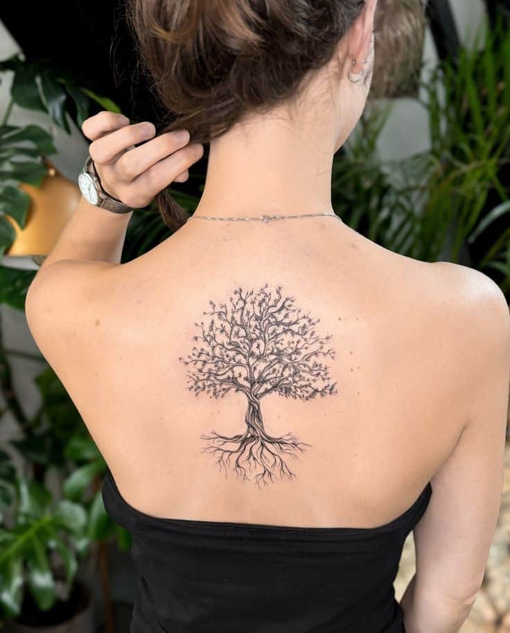 Woman with a detailed tree tattoo on her back, wearing a black top, indoors with plants in the background.