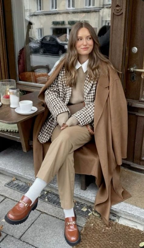 Woman in chic fall outfit with a brown coat and loafers, sitting at a café table outside with a cup of coffee.