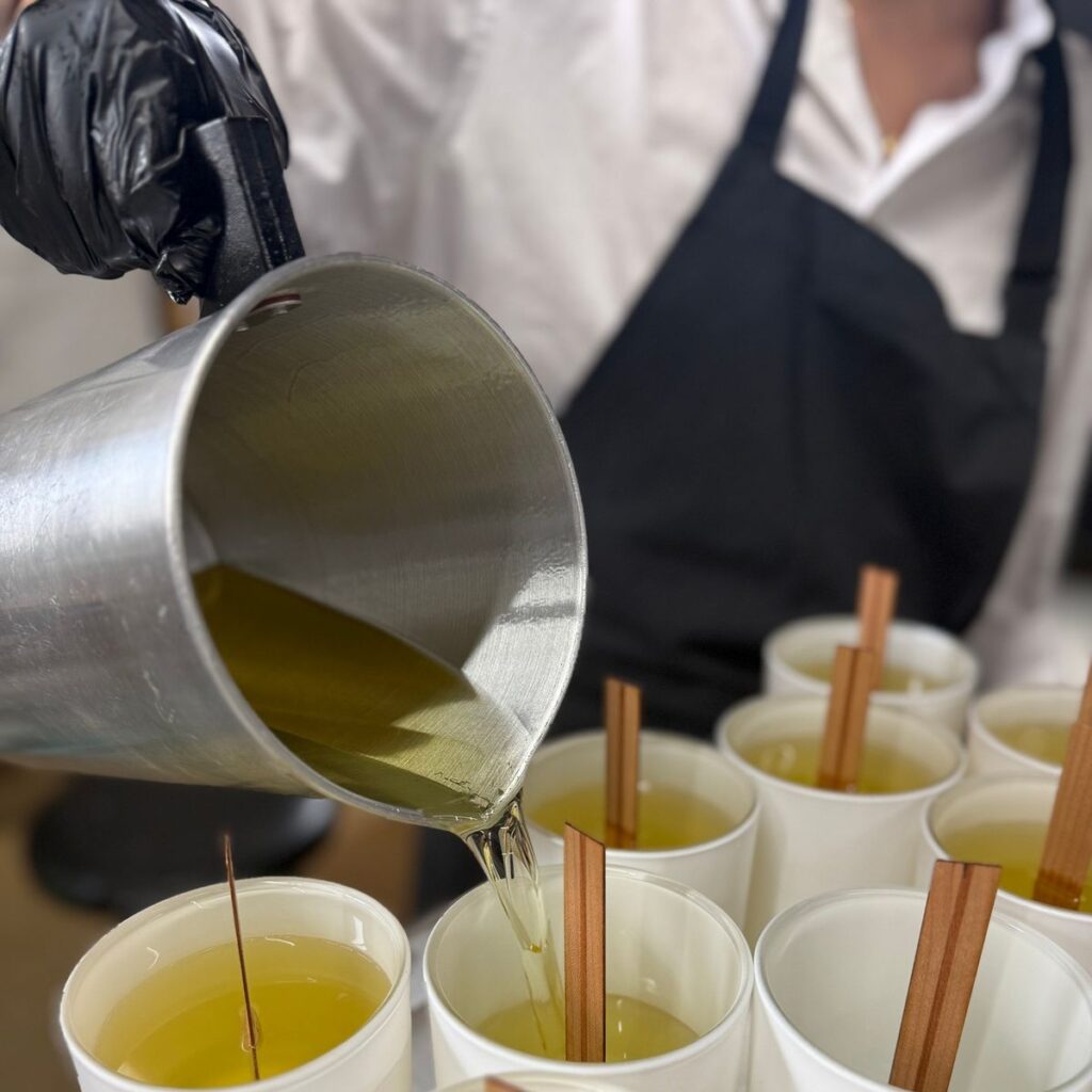 428b6efa50b5e9189a7771423ee08f5c Person pouring hot wax into white candle molds with wooden wicks, wearing a black apron and gloves. | Sky Rye Design Person pouring hot wax into white candle molds with wooden wicks, wearing a black apron and gloves.
